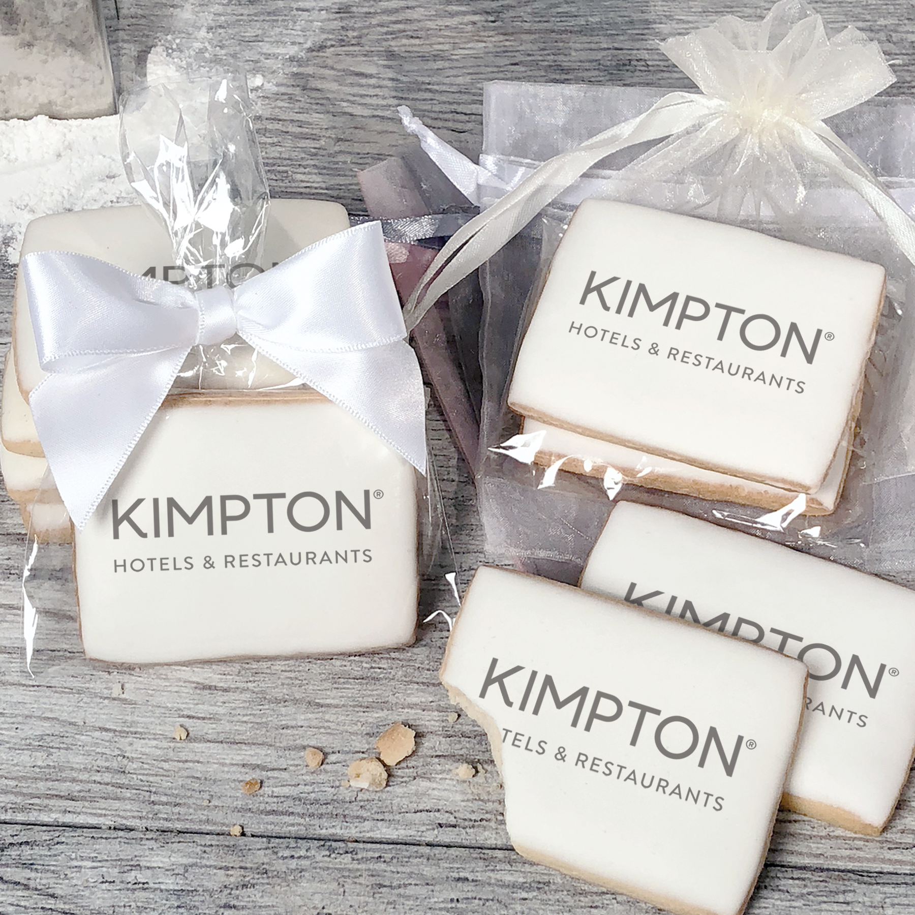 A stack of rectangular butter shortbread cookies with Kimpton logo printed directly on a white, light sugar icing. Some cookies are shown in clear packaging with a twist-tie ribbon bow or inside an organza bag.