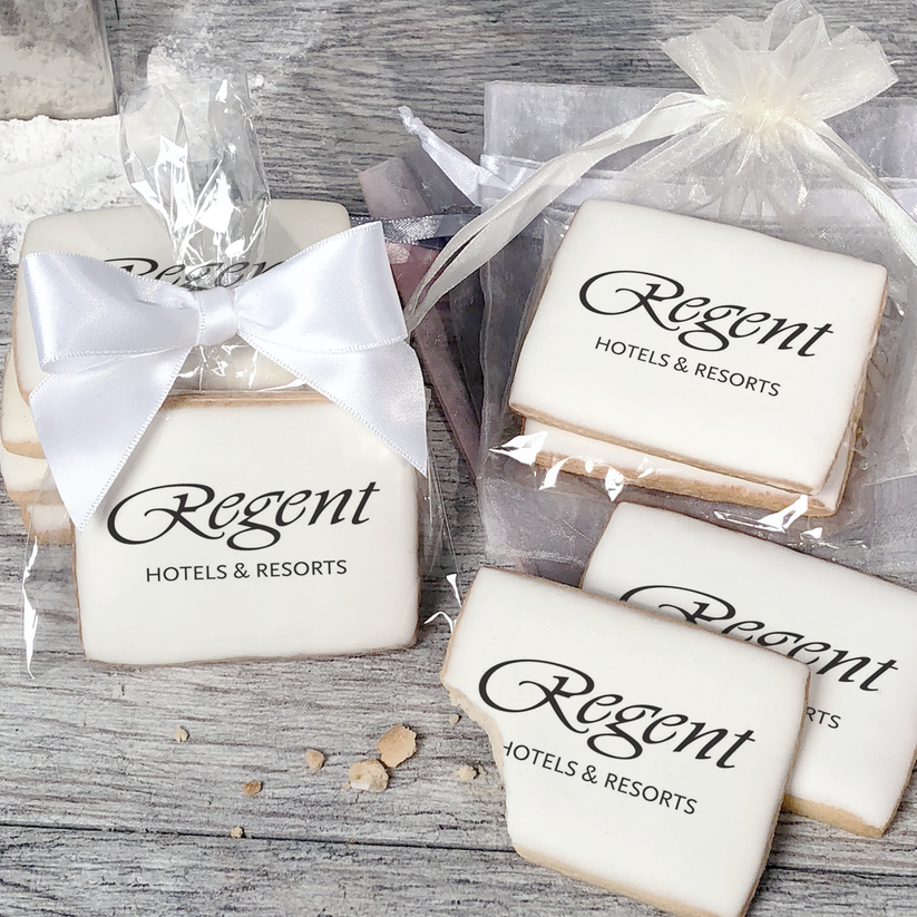 A stack of rectangular butter shortbread cookies with Regent logo printed directly on a white, light sugar icing. Some cookies are shown in clear packaging with a twist-tie ribbon bow or inside an organza bag.