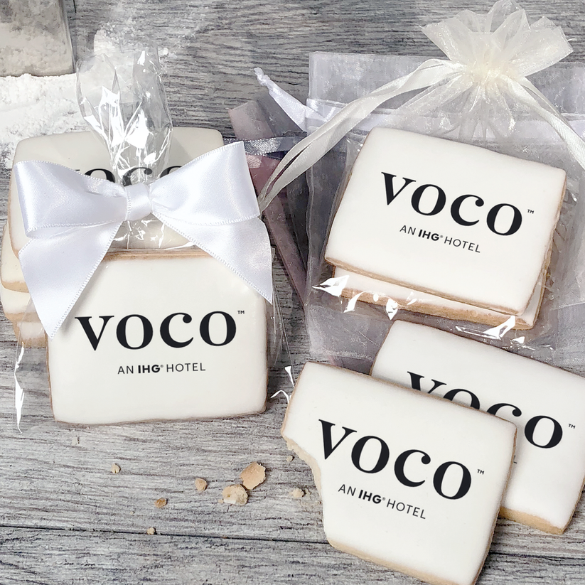 A stack of rectangular butter shortbread cookies with Voco logo printed directly on a white, light sugar icing. Some cookies are shown in clear packaging with a twist-tie ribbon bow or inside an organza bag.