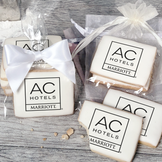 A stack of rectangular butter shortbread cookies with AC Hotels logo printed directly on a white, light sugar icing. Some cookies are shown in clear packaging with a twist-tie ribbon bow or inside an organza bag.