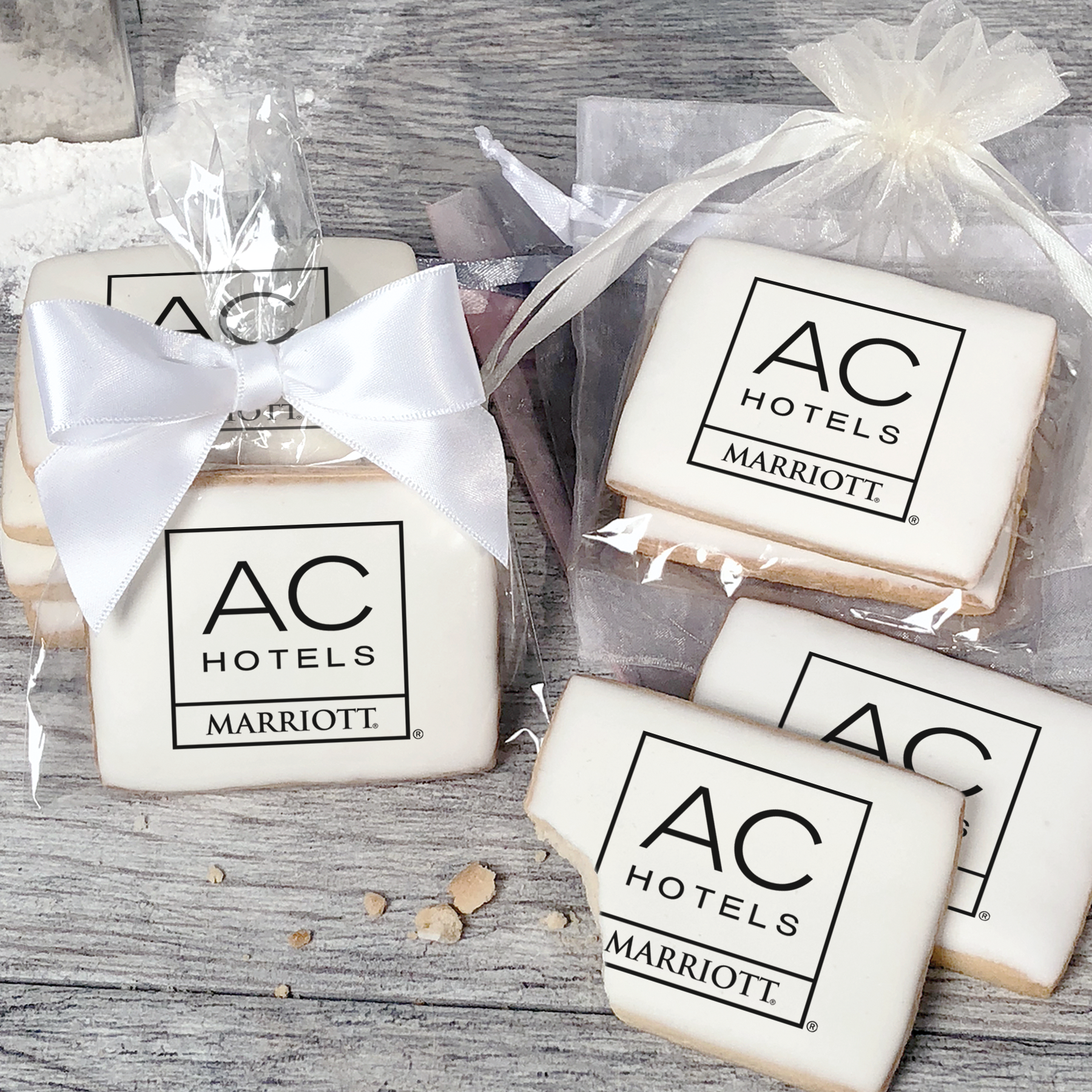 A stack of rectangular butter shortbread cookies with AC Hotels logo printed directly on a white, light sugar icing. Some cookies are shown in clear packaging with a twist-tie ribbon bow or inside an organza bag.