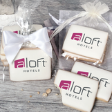 A stack of rectangular butter shortbread cookies with Aloft Hotels logo printed directly on a white, light sugar icing. Some cookies are shown in clear packaging with a twist-tie ribbon bow or inside an organza bag.