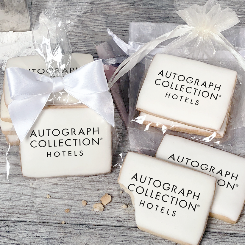 A stack of rectangular butter shortbread cookies with Autograph Collection logo printed directly on a white, light sugar icing. Some cookies are shown in clear packaging with a twist-tie ribbon bow or inside an organza bag.