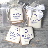 A stack of rectangular butter shortbread cookies with Delta Hotels logo printed directly on a white, light sugar icing. Some cookies are shown in clear packaging with a twist-tie ribbon bow or inside an organza bag.
