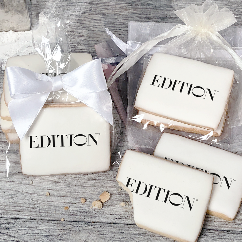 A stack of rectangular butter shortbread cookies with Edition logo printed directly on a white, light sugar icing. Some cookies are shown in clear packaging with a twist-tie ribbon bow or inside an organza bag.