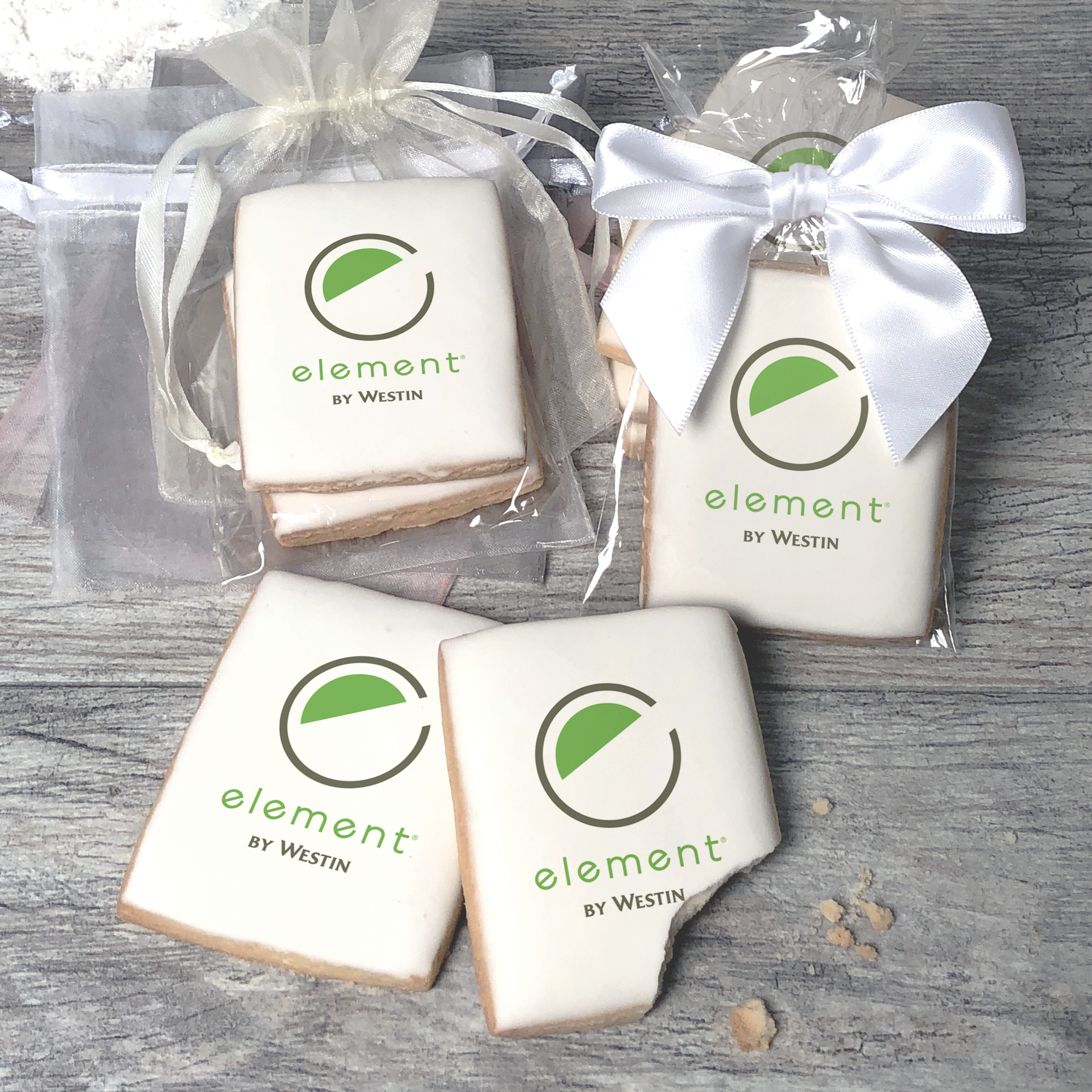 A stack of rectangular butter shortbread cookies with Element by Westin logo printed directly on a white, light sugar icing. Some cookies are shown in clear packaging with a twist-tie ribbon bow or inside an organza bag.