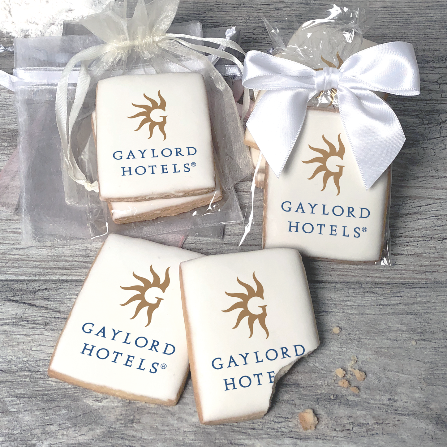 A stack of rectangular butter shortbread cookies with Gaylord Hotels logo printed directly on a white, light sugar icing. Some cookies are shown in clear packaging with a twist-tie ribbon bow or inside an organza bag.