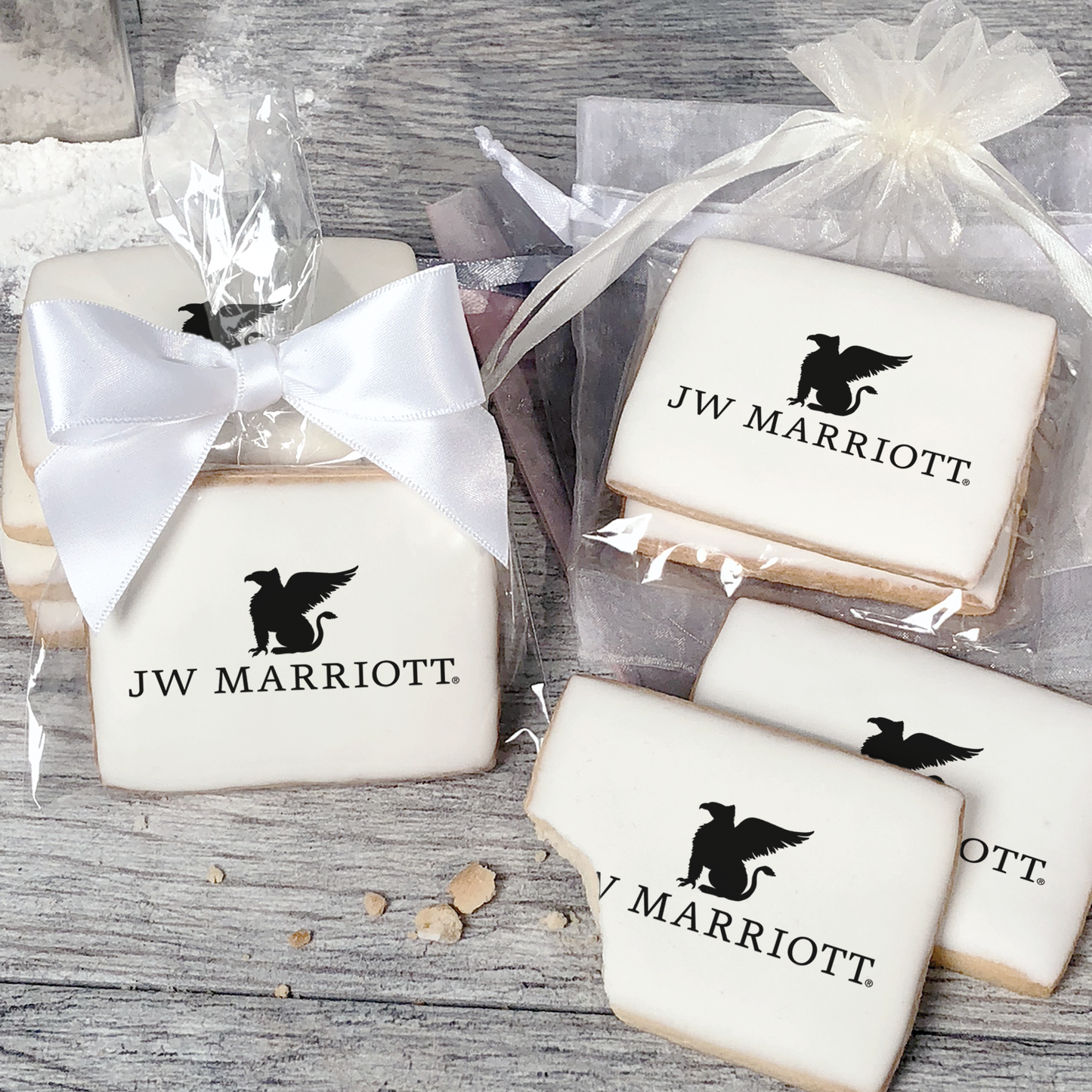 A stack of rectangular butter shortbread cookies with JW Marriott logo printed directly on a white, light sugar icing. Some cookies are shown in clear packaging with a twist-tie ribbon bow or inside an organza bag.