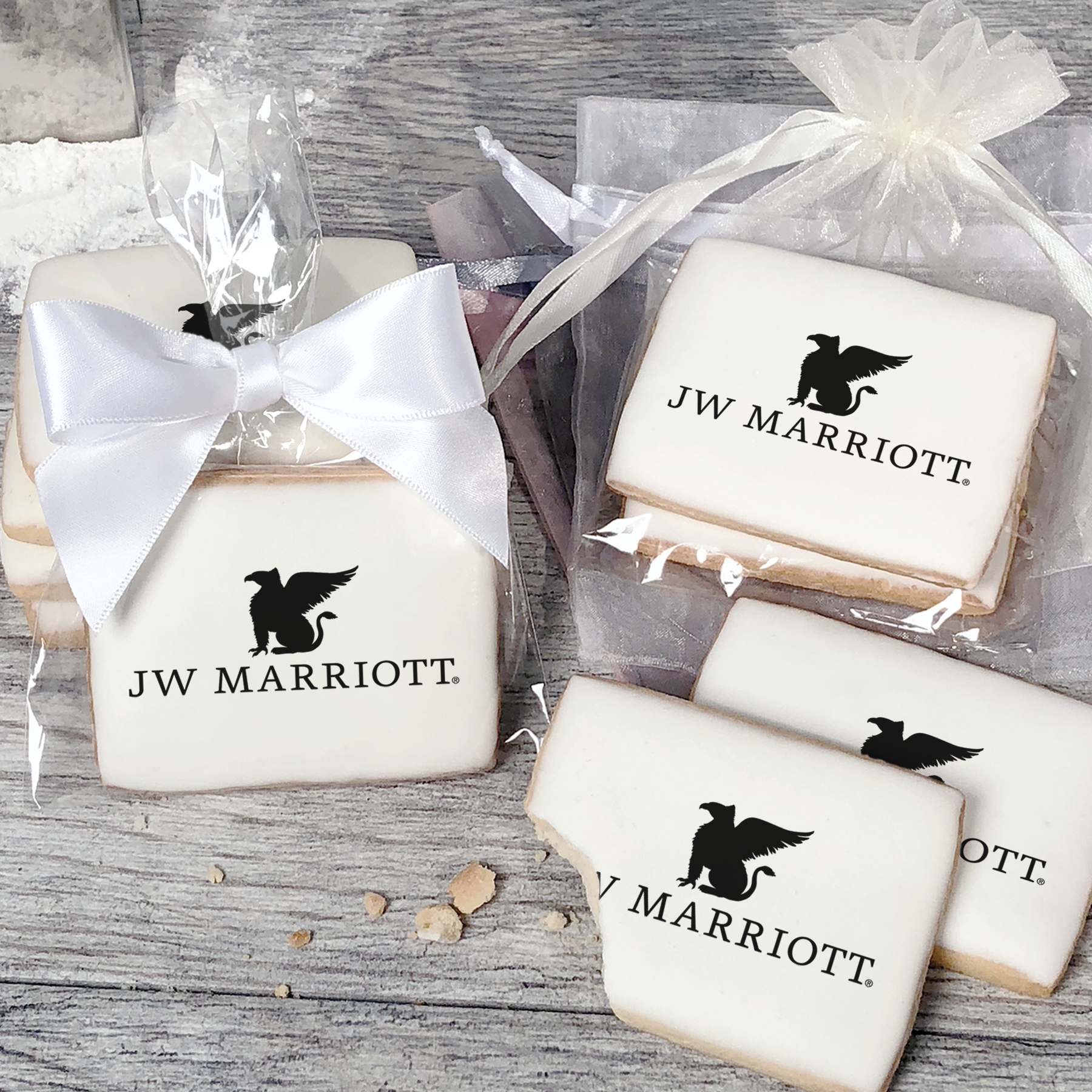 A stack of rectangular butter shortbread cookies with JW Marriott logo printed directly on a white, light sugar icing. Some cookies are shown in clear packaging with a twist-tie ribbon bow or inside an organza bag.