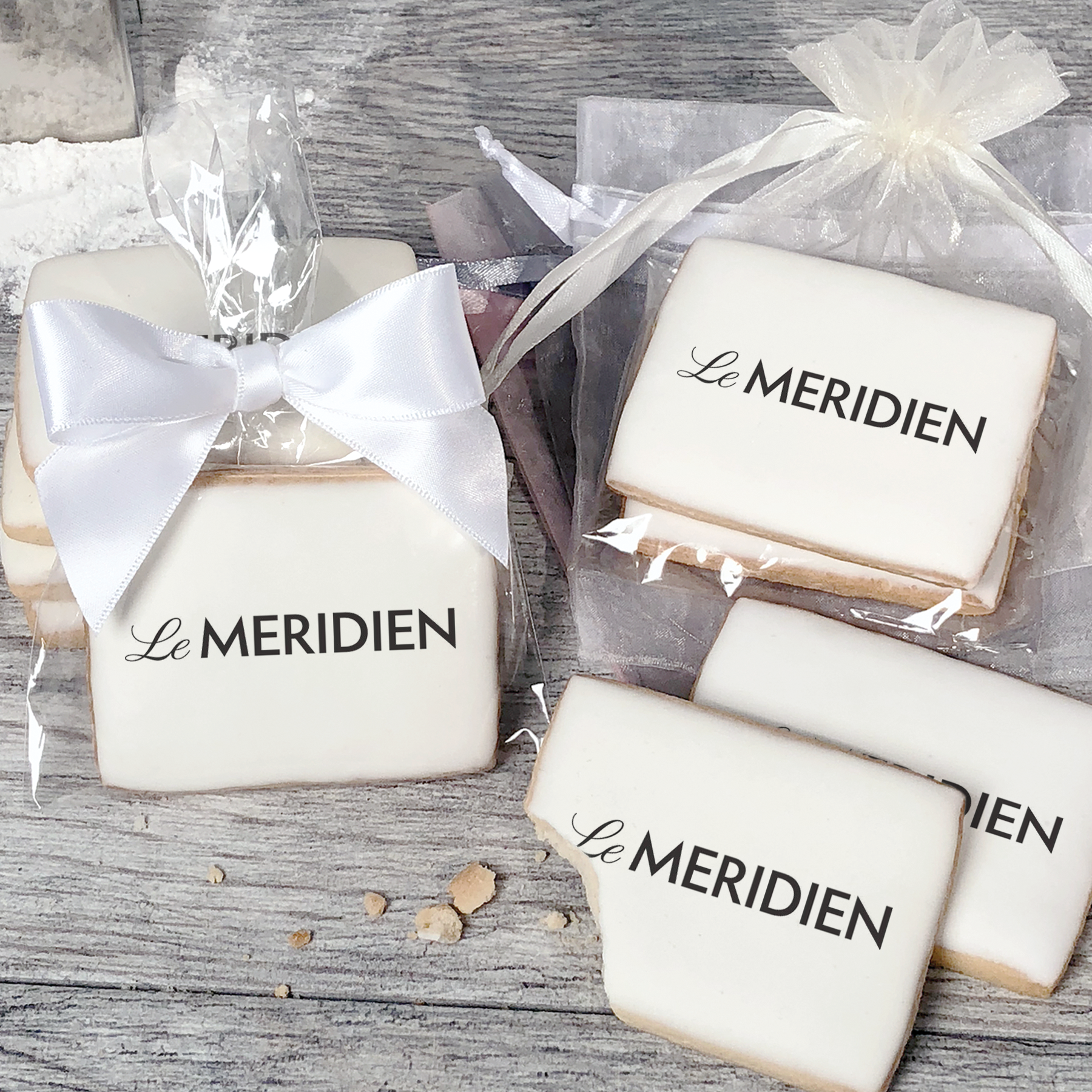 A stack of rectangular butter shortbread cookies with Le Meridien logo printed directly on a white, light sugar icing. Some cookies are shown in clear packaging with a twist-tie ribbon bow or inside an organza bag.
