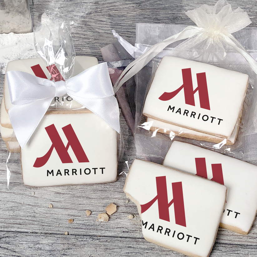 A stack of rectangular butter shortbread cookies with Marriott logo printed directly on a white, light sugar icing. Some cookies are shown in clear packaging with a twist-tie ribbon bow or inside an organza bag.