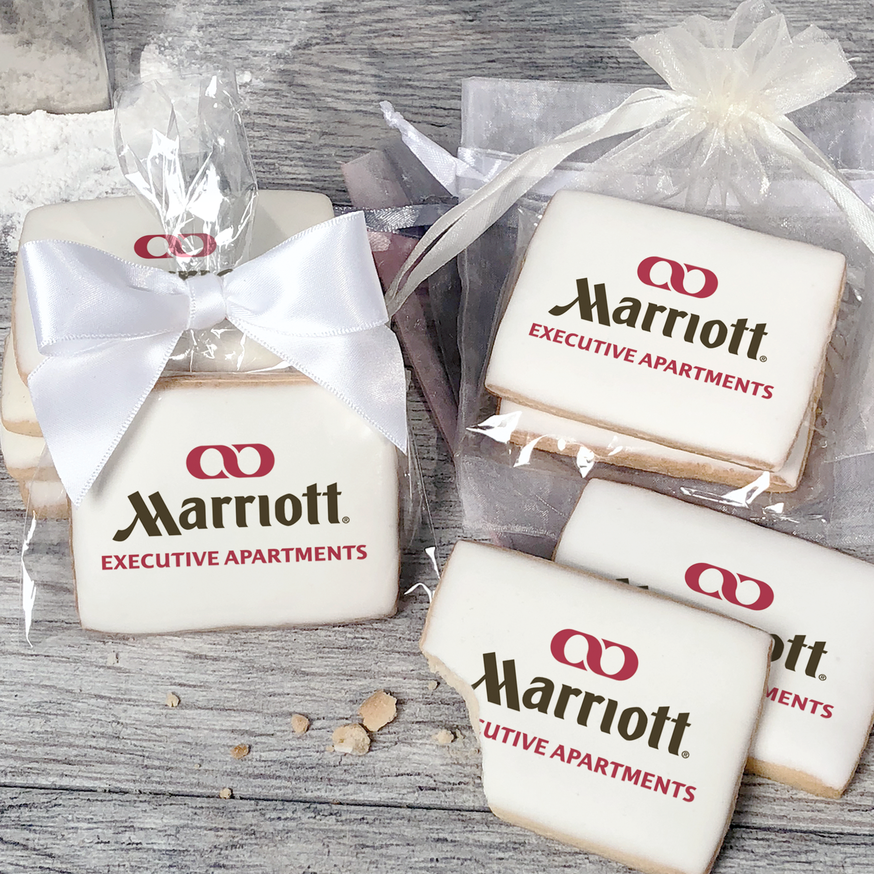 A stack of rectangular butter shortbread cookies with Marriott Executive Apartments logo printed directly on a white, light sugar icing. Some cookies are shown in clear packaging with a twist-tie ribbon bow or inside an organza bag.