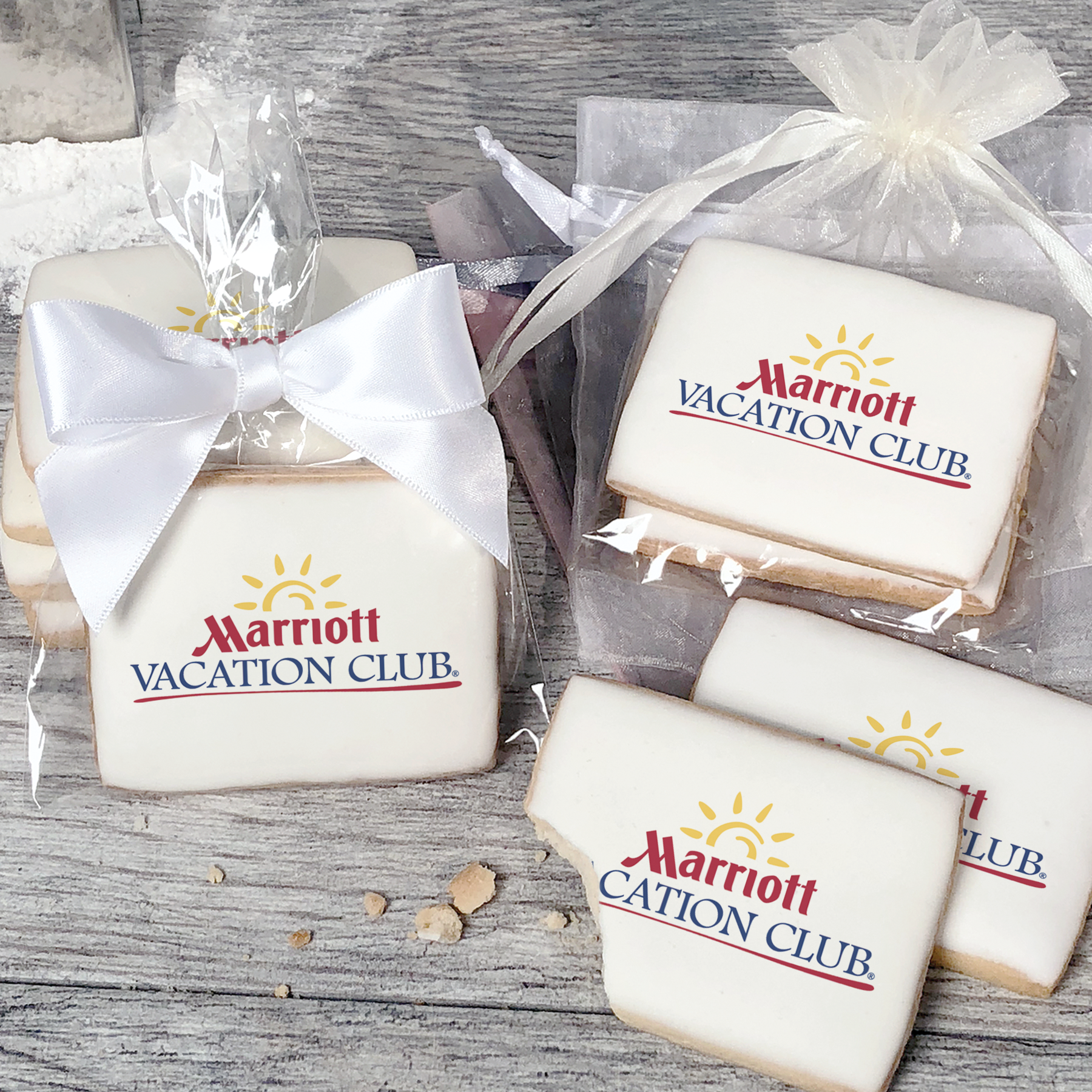 A stack of rectangular butter shortbread cookies with Marriott Vacation Club logo printed directly on a white, light sugar icing. Some cookies are shown in clear packaging with a twist-tie ribbon bow or inside an organza bag.