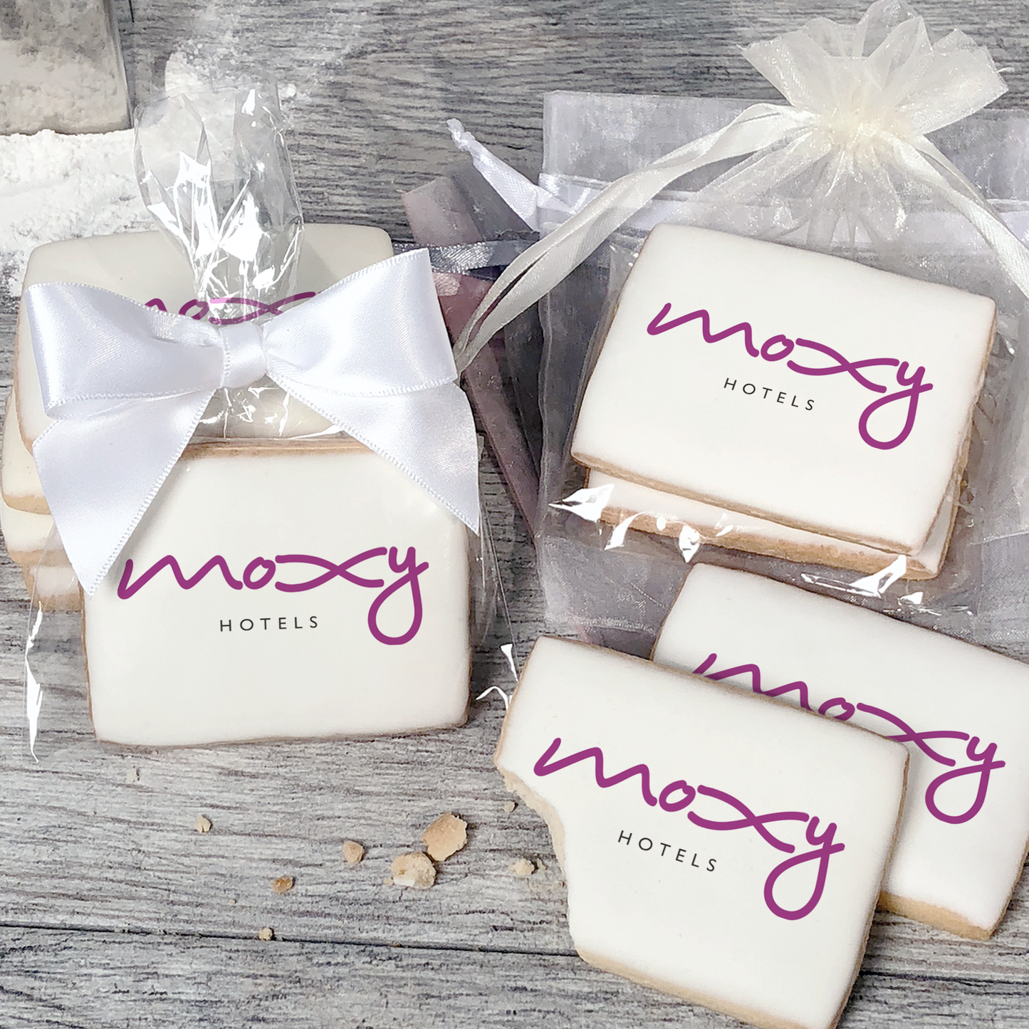 A stack of rectangular butter shortbread cookies with Moxy Hotels logo printed directly on a white, light sugar icing. Some cookies are shown in clear packaging with a twist-tie ribbon bow or inside an organza bag.