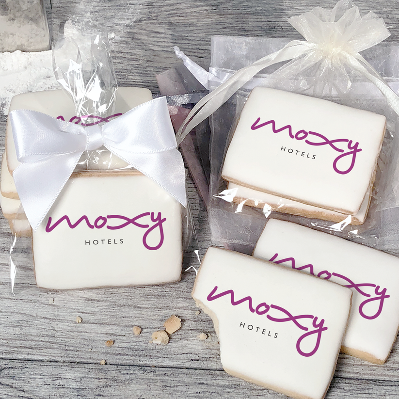 A stack of rectangular butter shortbread cookies with Moxy Hotels logo printed directly on a white, light sugar icing. Some cookies are shown in clear packaging with a twist-tie ribbon bow or inside an organza bag.