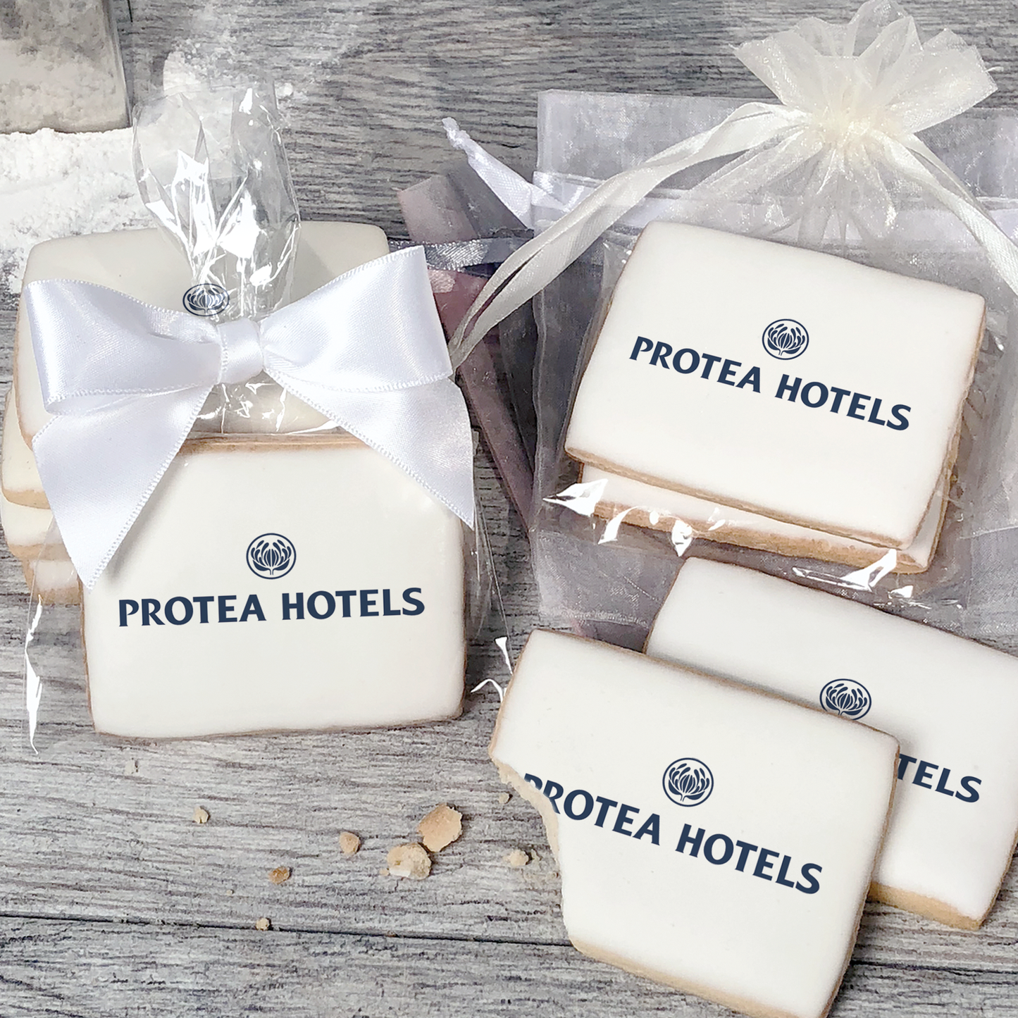 A stack of rectangular butter shortbread cookies with Protea Hotels logo printed directly on a white, light sugar icing. Some cookies are shown in clear packaging with a twist-tie ribbon bow or inside an organza bag.