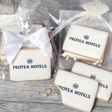 A stack of rectangular butter shortbread cookies with Protea Hotels logo printed directly on a white, light sugar icing. Some cookies are shown in clear packaging with a twist-tie ribbon bow or inside an organza bag.