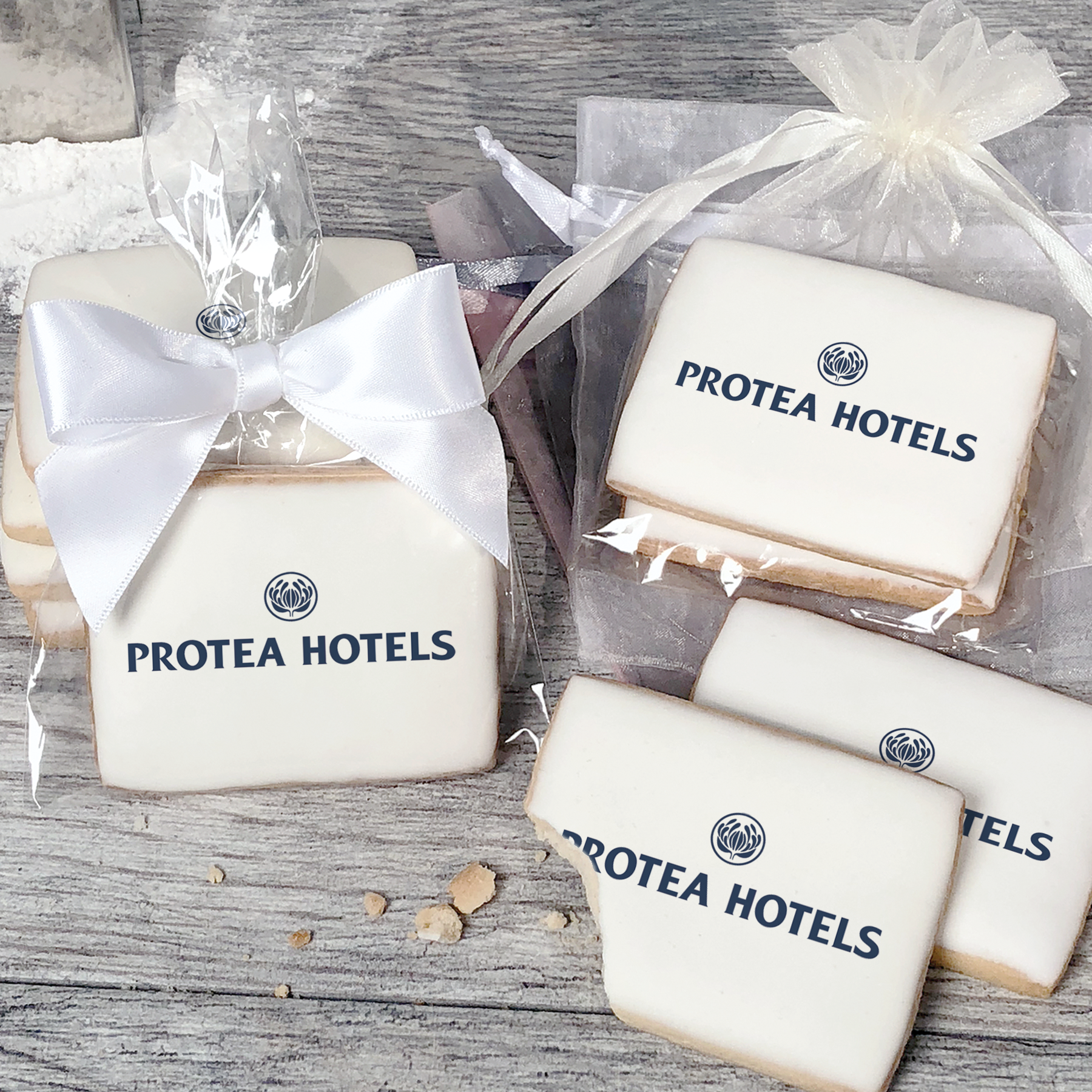 A stack of rectangular butter shortbread cookies with Protea Hotels logo printed directly on a white, light sugar icing. Some cookies are shown in clear packaging with a twist-tie ribbon bow or inside an organza bag.
