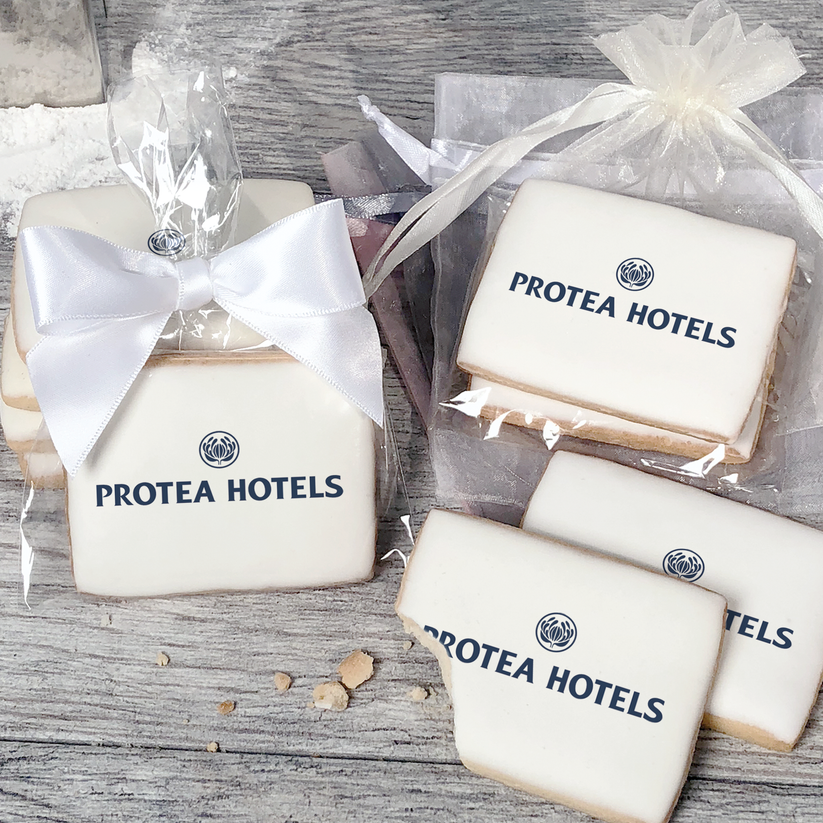 A stack of rectangular butter shortbread cookies with Protea Hotels logo printed directly on a white, light sugar icing. Some cookies are shown in clear packaging with a twist-tie ribbon bow or inside an organza bag.