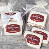A stack of rectangular butter shortbread cookies with Residence Inn logo printed directly on a white, light sugar icing. Some cookies are shown in clear packaging with a twist-tie ribbon bow or inside an organza bag.