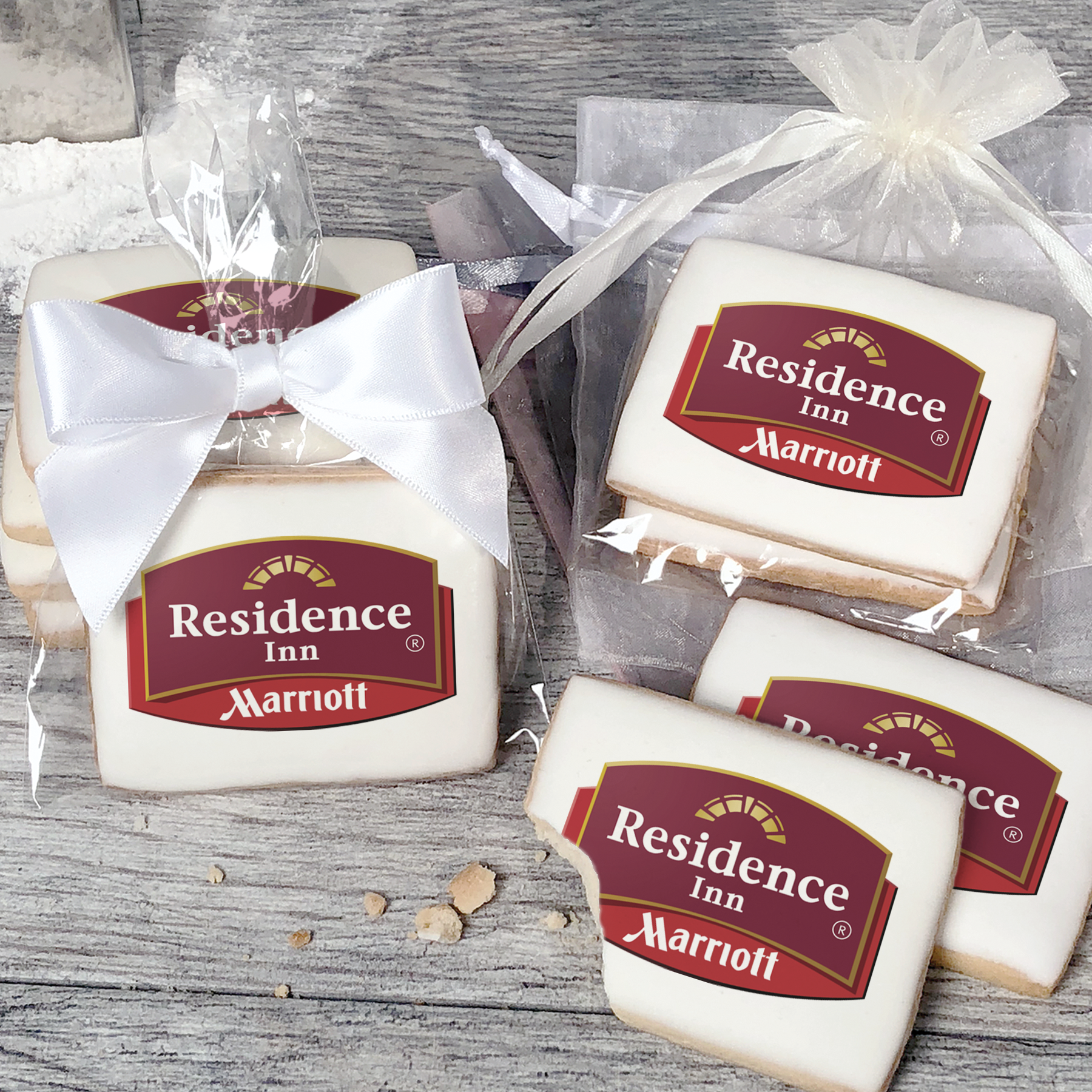 A stack of rectangular butter shortbread cookies with Residence Inn logo printed directly on a white, light sugar icing. Some cookies are shown in clear packaging with a twist-tie ribbon bow or inside an organza bag.
