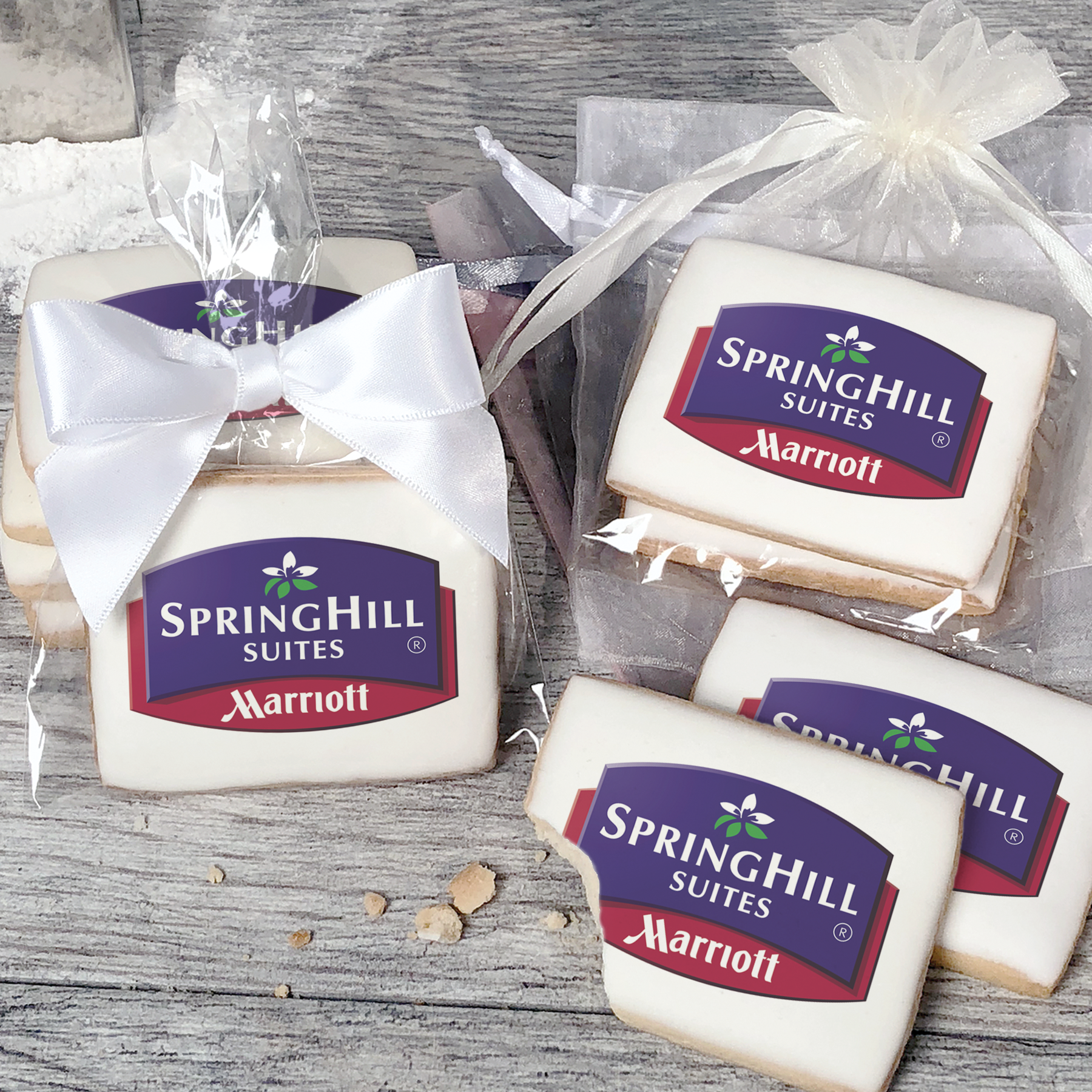 A stack of rectangular butter shortbread cookies with Springhill Suites logo printed directly on a white, light sugar icing. Some cookies are shown in clear packaging with a twist-tie ribbon bow or inside an organza bag.
