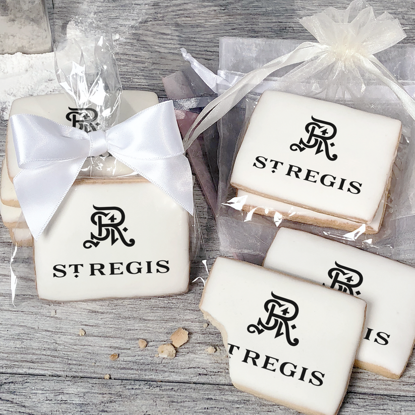 A stack of rectangular butter shortbread cookies with St Regis logo printed directly on a white, light sugar icing. Some cookies are shown in clear packaging with a twist-tie ribbon bow or inside an organza bag.