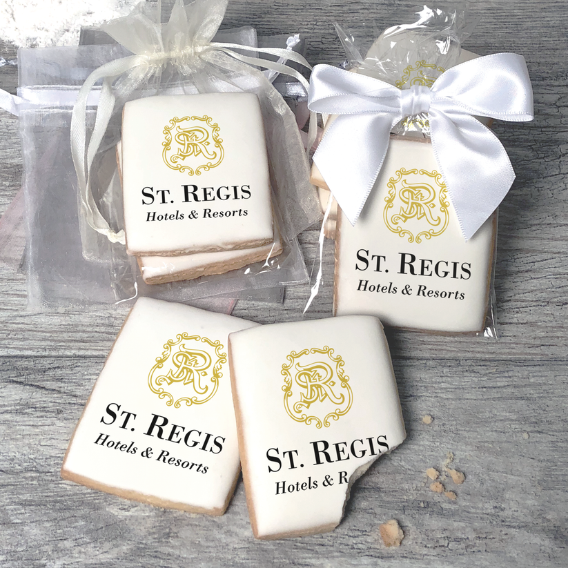 A stack of rectangular butter shortbread cookies with St Regis Hotels & Resorts logo printed directly on a white, light sugar icing. Some cookies are shown in clear packaging with a twist-tie ribbon bow or inside an organza bag.
