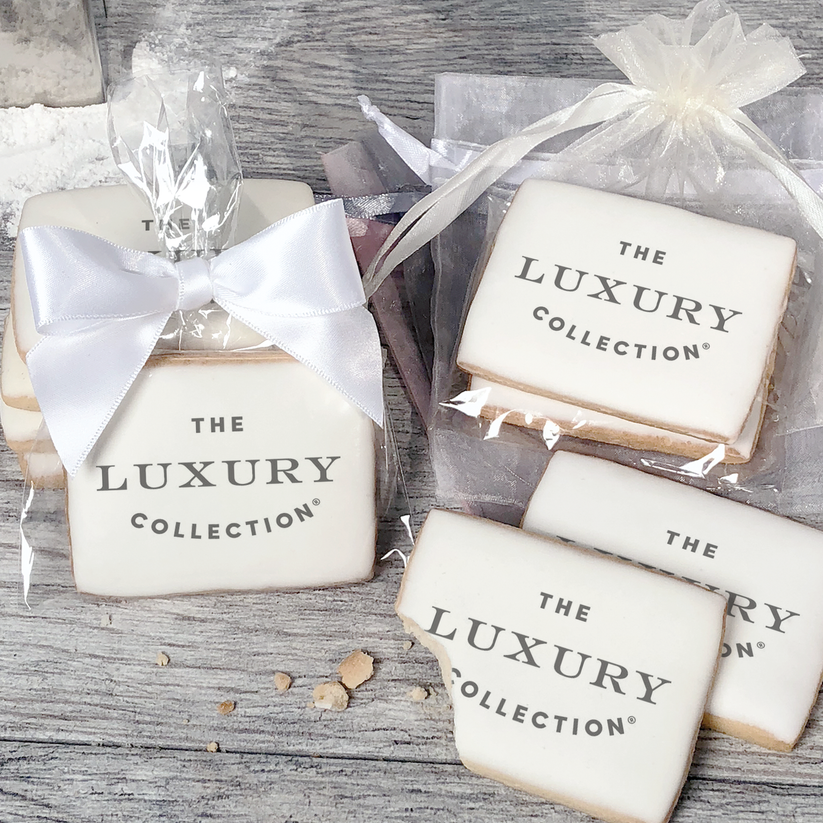 A stack of rectangular butter shortbread cookies with The Luxury Collection logo printed directly on a white, light sugar icing. Some cookies are shown in clear packaging with a twist-tie ribbon bow or inside an organza bag.