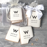 A stack of rectangular butter shortbread cookies with W Hotels logo printed directly on a white, light sugar icing. Some cookies are shown in clear packaging with a twist-tie ribbon bow or inside an organza bag.