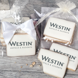 A stack of rectangular butter shortbread cookies with Westin Hotel & Resorts logo printed directly on a white, light sugar icing. Some cookies are shown in clear packaging with a twist-tie ribbon bow or inside an organza bag.