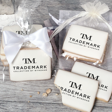 Load image into Gallery viewer, A stack of rectangular butter shortbread cookies with TM Trademark Collection by Wyndham logo printed directly on a white, light sugar icing. Some cookies are shown in clear packaging with a twist-tie ribbon bow or inside an organza bag.