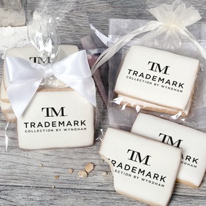 A stack of rectangular butter shortbread cookies with TM Trademark Collection by Wyndham logo printed directly on a white, light sugar icing. Some cookies are shown in clear packaging with a twist-tie ribbon bow or inside an organza bag.