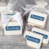 A stack of rectangular butter shortbread cookies with Travelodge logo printed directly on a white, light sugar icing. Some cookies are shown in clear packaging with a twist-tie ribbon bow or inside an organza bag.