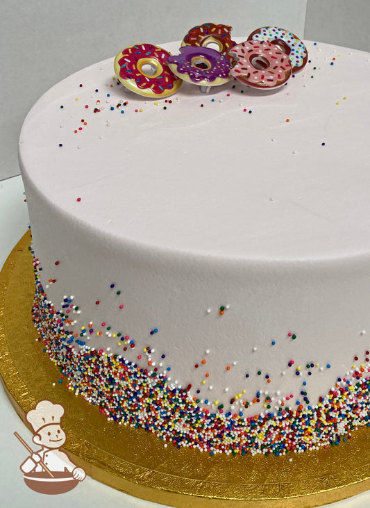 Single tier cake decorated with multi-color sprinkles on the bottom half of the cake wall and some sprinkled on top and plastic donut rings.