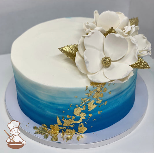 Cake with white icing, decorated with blue-watercolor, sugar magnolia flowers with gold accents and gold foil cascading on the front of the cake.
