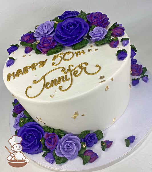 Single tier cake with white icing and decorated with buttercream roses in purple colors, gold writing and gold foil flakes all around the cake.