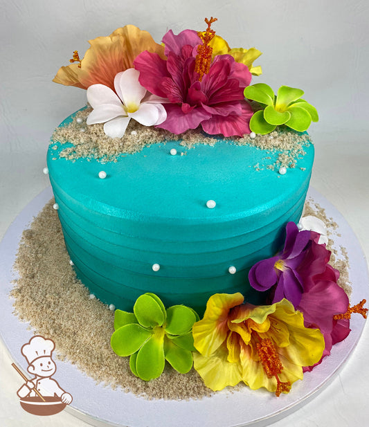 Single tier cake with teal colored icing and decorated with a horizontal texture, multi-color silk Hawaiian flowers and sugar sand.