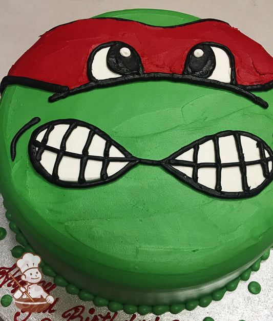 Single tier cake with green icing, decorated with a hand-piped face to look like Raphael from the Ninja Turtles.