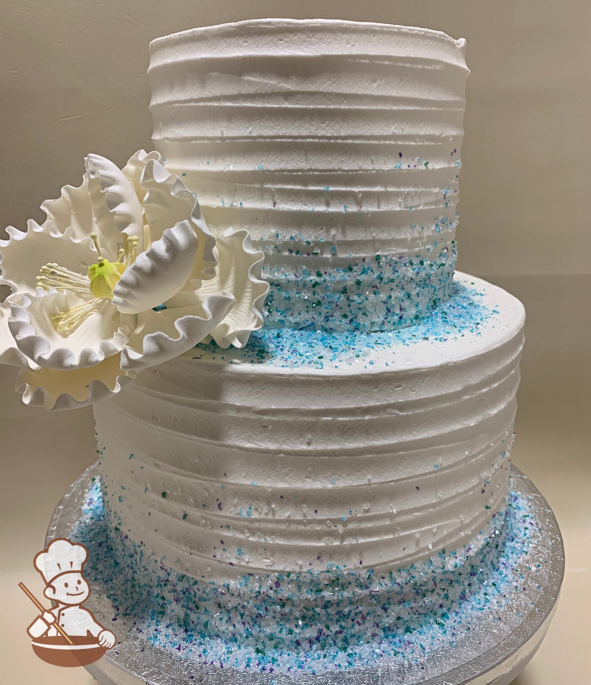 2-tier cake with white icing in a horizontal texture pattern, with blue sugar crystal on the base of the tiers and one white sugar peony flower.