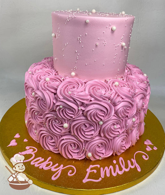2-tier cake with pink buttercream rosettes on the bottom tier and smooth pink icing on the top tier, the whole cake is covered with white pearls.