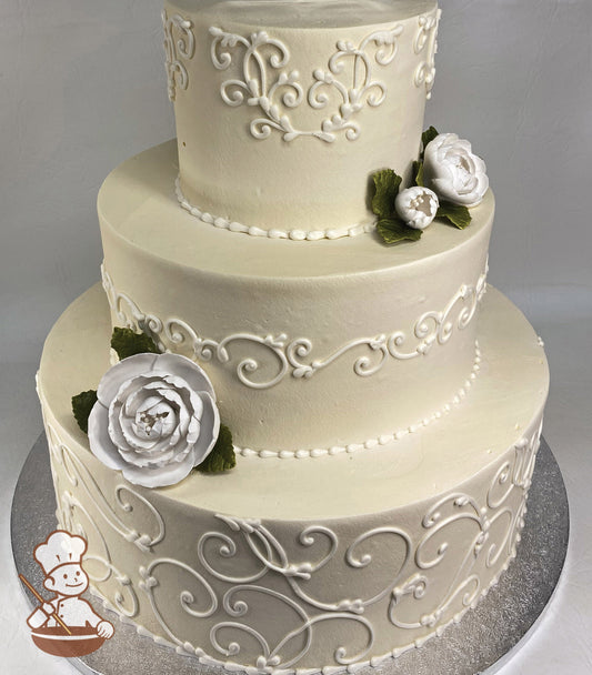 3-tier round cake with ivory icing and white buttercream scrolls on all tiers. The cake also has white sugar flowers and green fondant leaves.