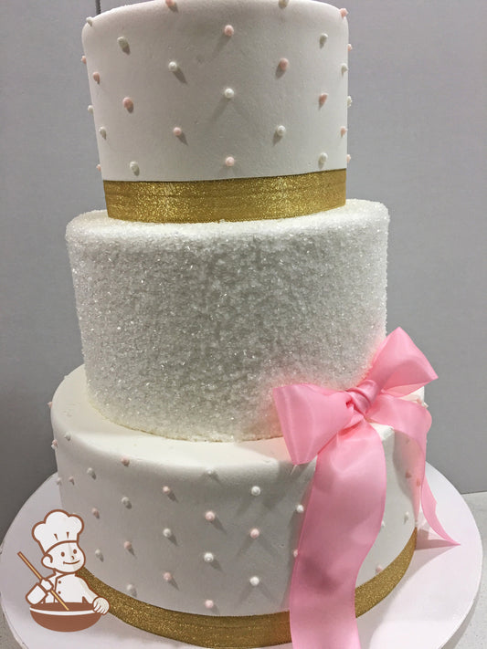 3-tier cake with white icing on the bottom and top tier with dots and a gold ribbon. The middle tier has white sugar crystal and a pink bow.