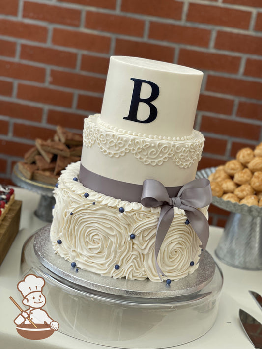 3-tier cake with a white ruffle bottom tier, white scrolls in the middle tier with a gray ribbon bow and a custom fondant letter on the top tier.