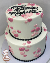 Two-tier round cake with white icing and buttercream hearts in shades of pink all over both tiers and black bead trims on bottoms of tiers.