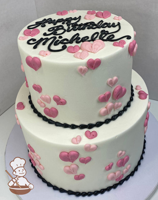Two-tier round cake with white icing and buttercream hearts in shades of pink all over both tiers and black bead trims on bottoms of tiers.