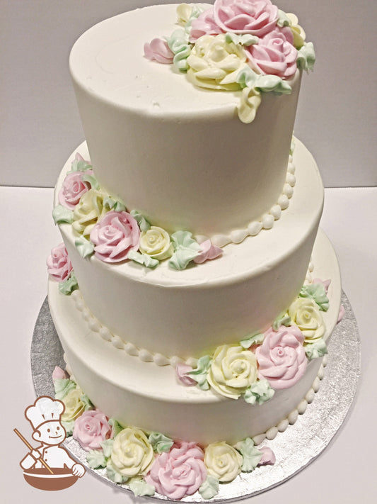 Three-tier round cake with pink and yellow buttercream roses, has green leaves and rosebuds with white bead trims on each bottom of the tiers.