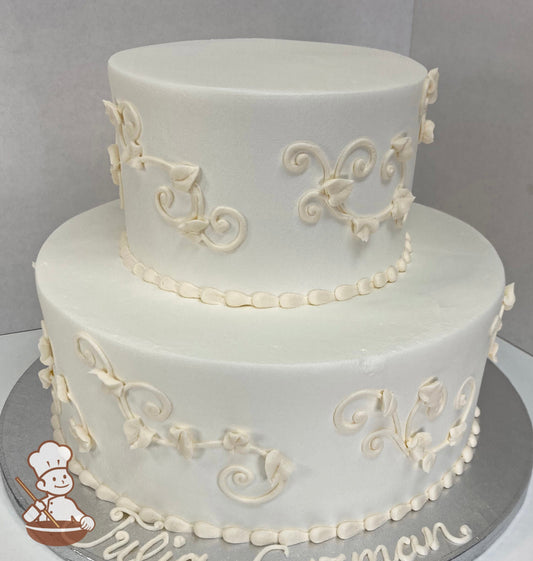 2-tier round cake with white icing, decorated with ivory-colored buttercream scrolls on cake walls with a beaded trim.