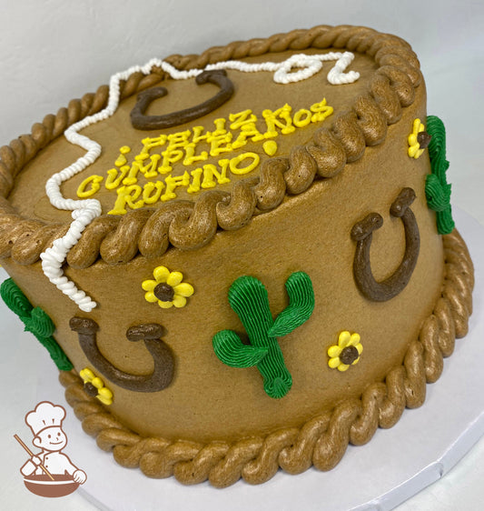 Round cake with brown icing, has western buttercream decoration including cactus, horseshoes and daisies on cake wall with rope trims.