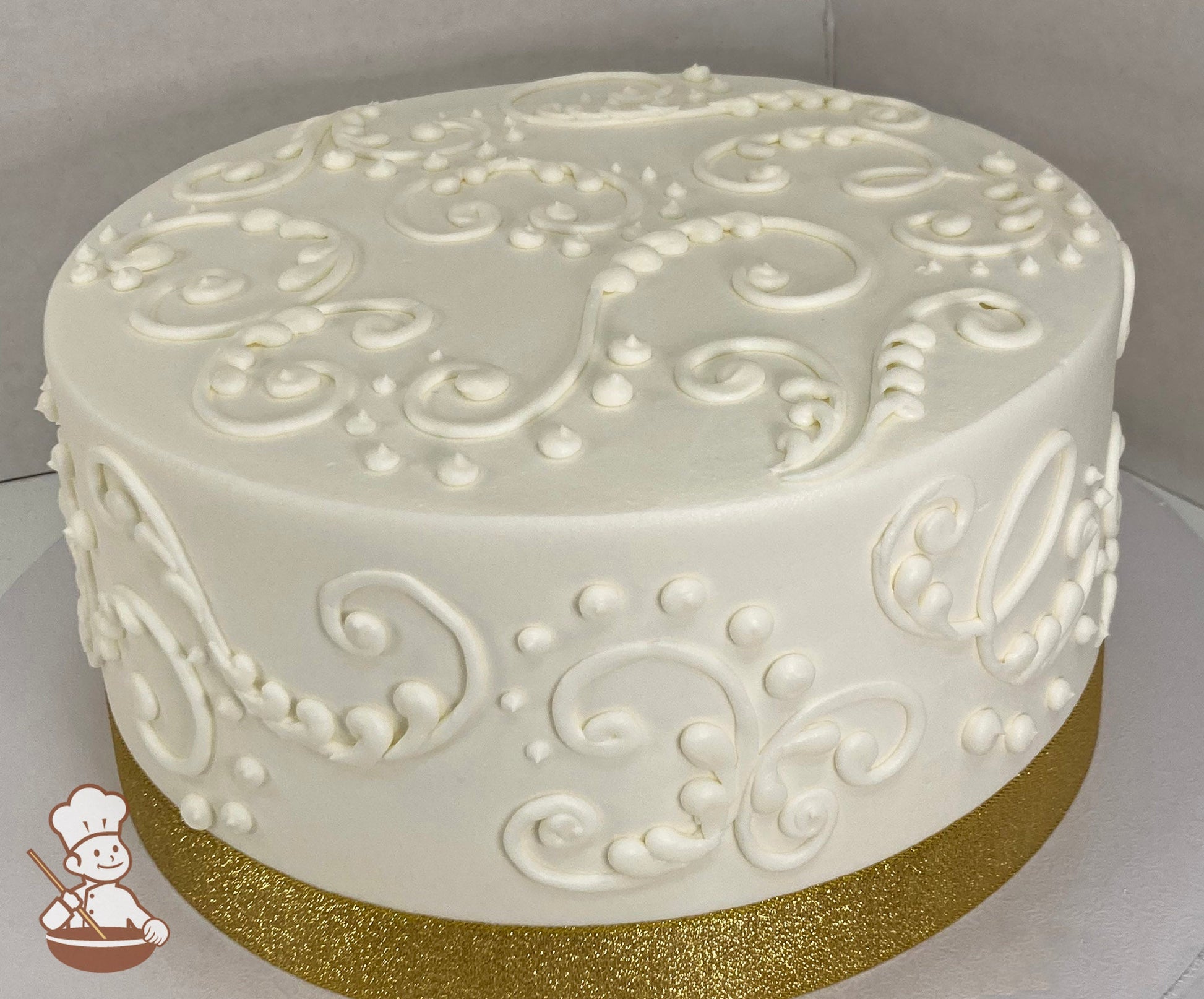 Round cake with white icing and white buttercream scrolls and dots on cake walls and on top of cake and a gold ribbon on the base of the cake.